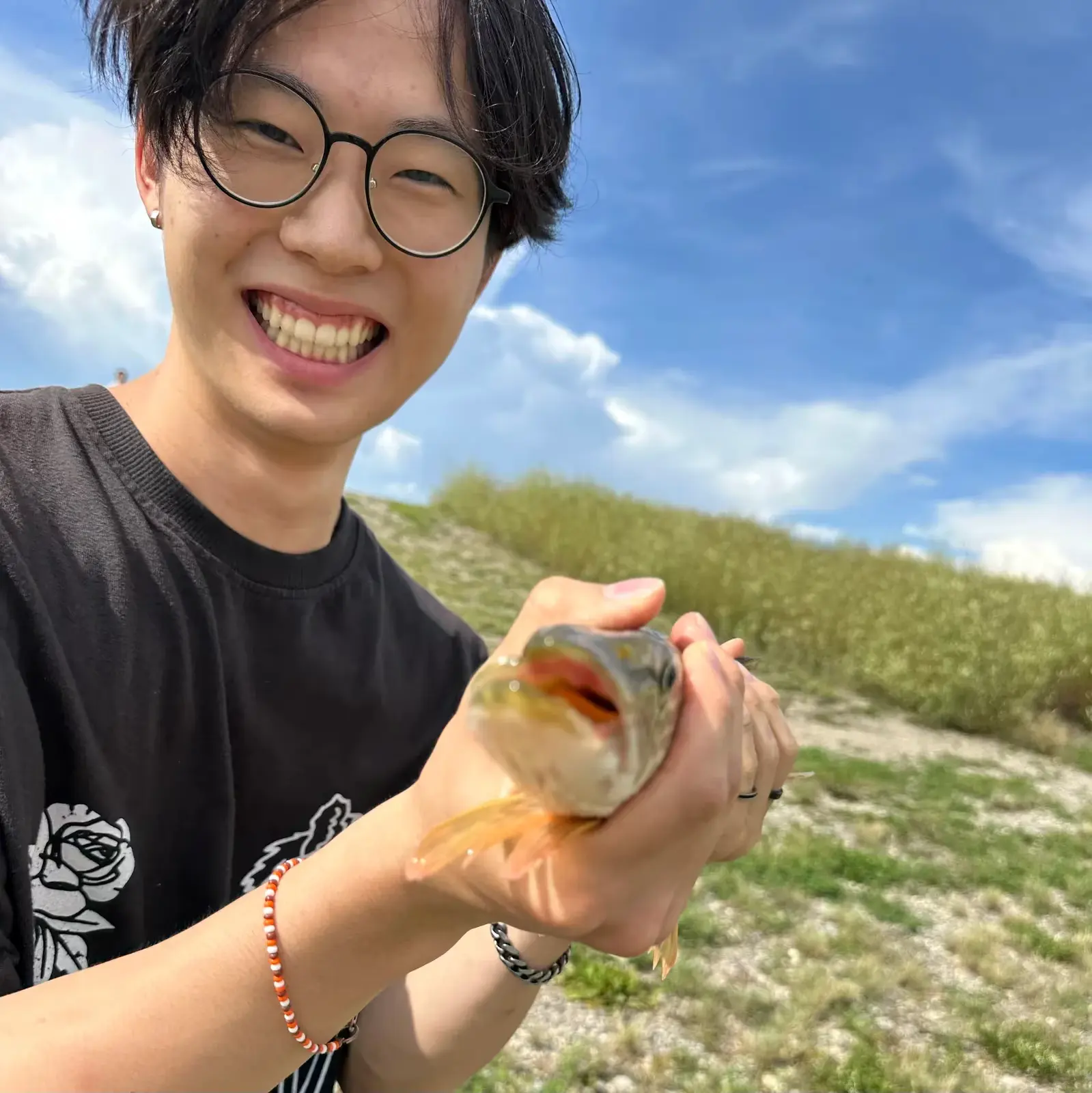 Liam smiling and holding a small fish he caught, with grassy hills and a blue sky behind him.